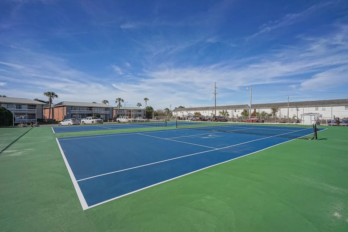 A vibrant tennis court with blue and green surfaces is featured in the center, surrounded by residential buildings. The setting is lively, inviting residents to play matches, hold casual games, or gather for community tournaments.