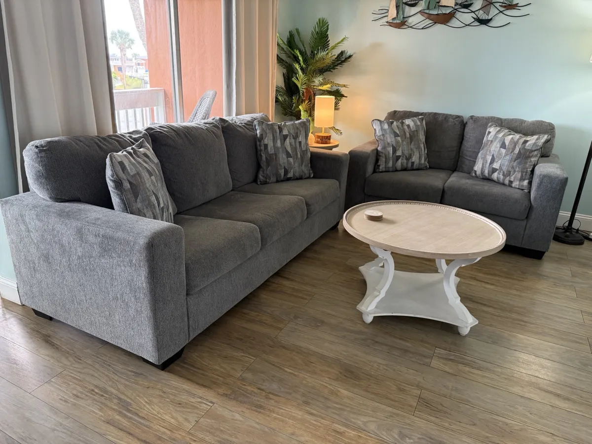 A welcoming living area with gray sofas, decorative pillows, a round coffee table, and lush indoor plants. Sunlight pours in, creating a cozy space ideal for both relaxing and working remotely.
