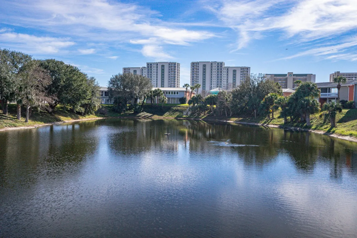 A serene lake stretches across the scene, bordered by lush greenery and overlooked by tall residential buildings. People enjoy quiet walks along the water and peaceful lakeside moments, perfect for picnicking, wildlife watching, or bringing pets along.