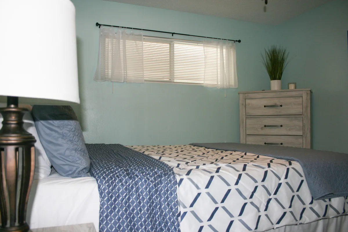 A serene queen bedroom featuring patterned bedding, a window adorned with sheer curtains letting in natural light, and a dresser with subtle, tasteful decor. The room exudes tranquility and comfort—perfect for restful nights or a peaceful morning start.