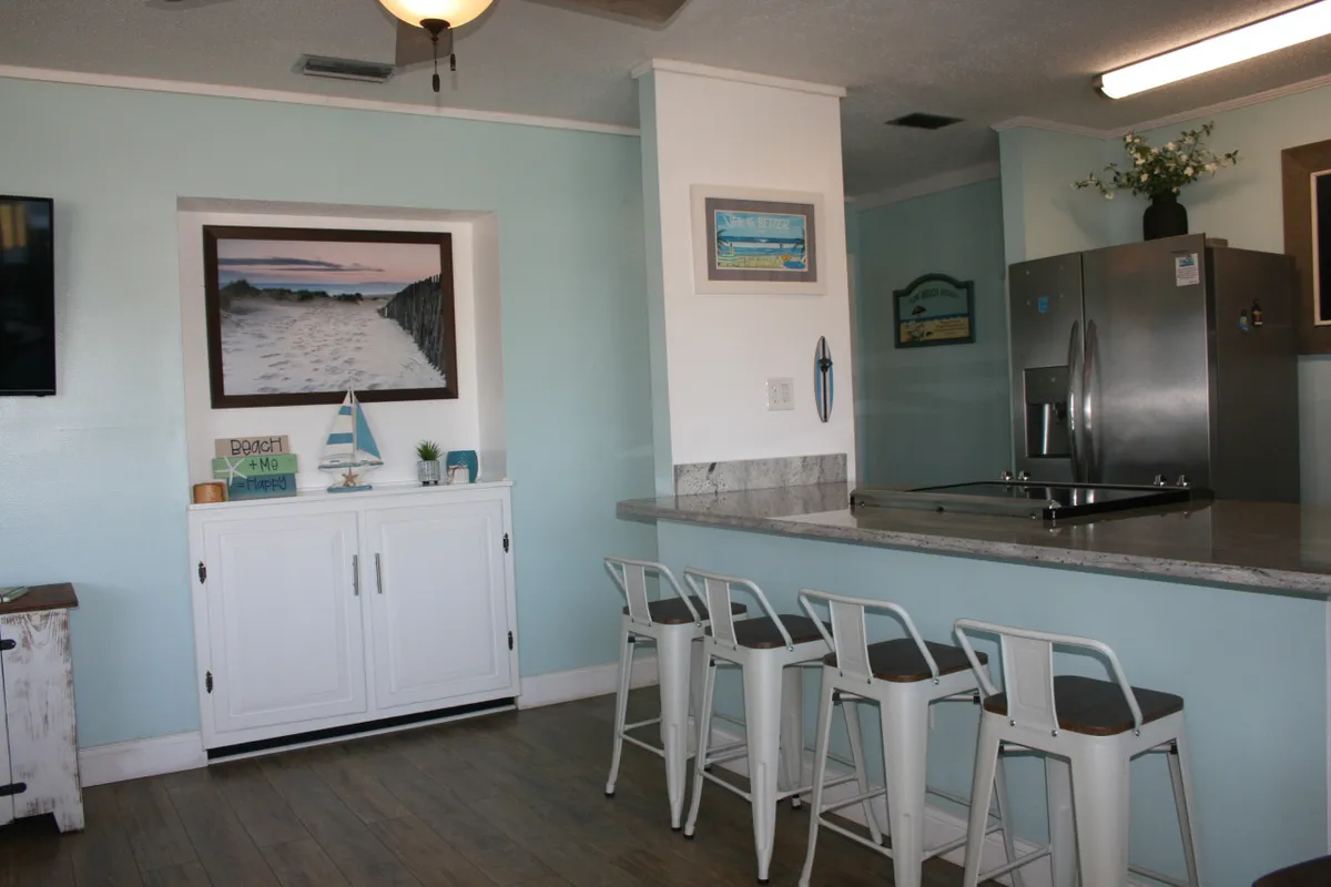 A bright, open kitchen area with white cabinetry, a central island with stools, and framed coastal artwork. The nautical elements and airy feel suggest a space meant for gathering and informal celebrations, reminiscent of time spent at a resort or on holiday.