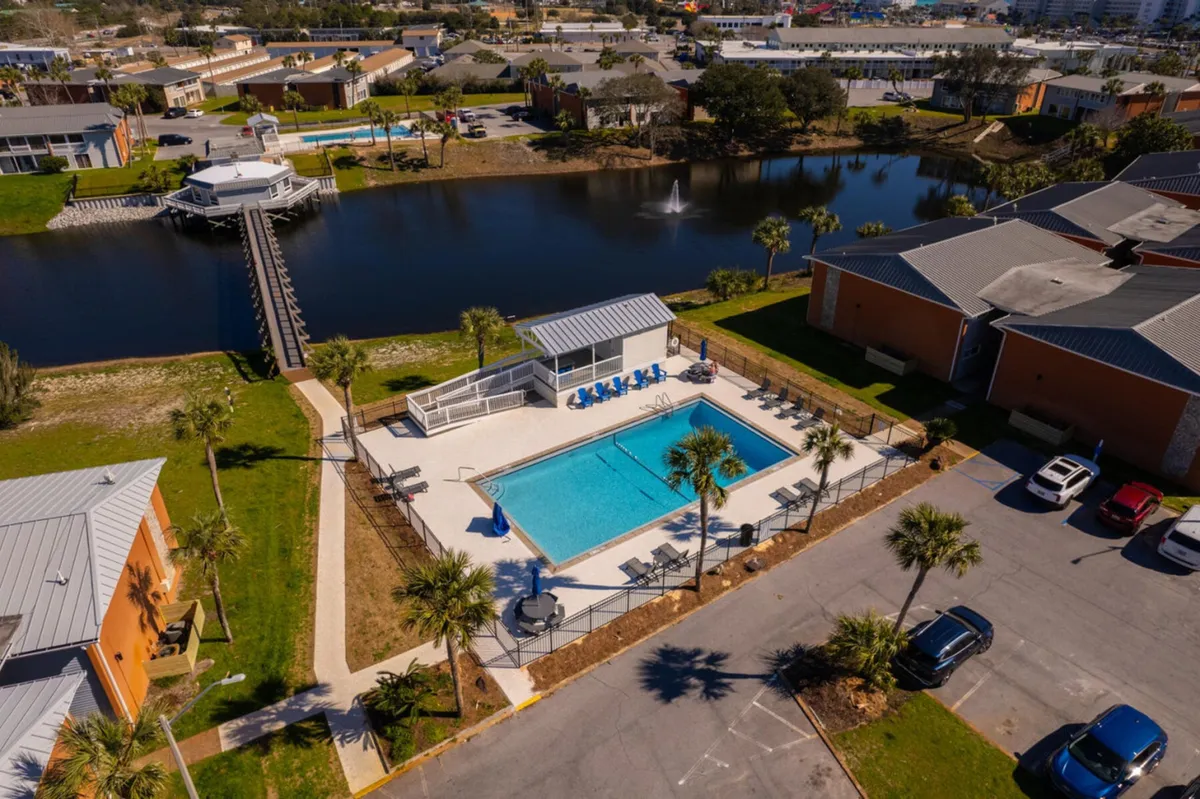 The hero image showcases a sparkling, rectangular swimming pool surrounded by inviting blue lounge chairs under a shaded pavilion. Beyond the pool, well-manicured pathways wind through lush palm trees and lead to a tranquil lake with a central fountain, all set within a vibrant, resort-style community.