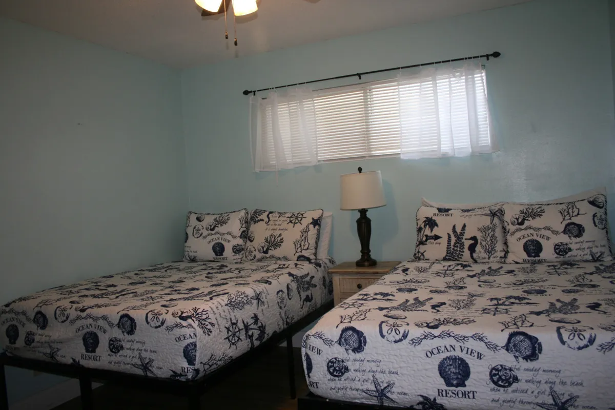 Two full beds with matching ocean-themed bedding are arranged side by side, separated by a central wooden nightstand with a lamp. Soft lighting and a window with white blinds and sheer curtains create a welcoming, peaceful vibe. The bedding and decor evoke a subtle coastal feel, ideal for families or friends.