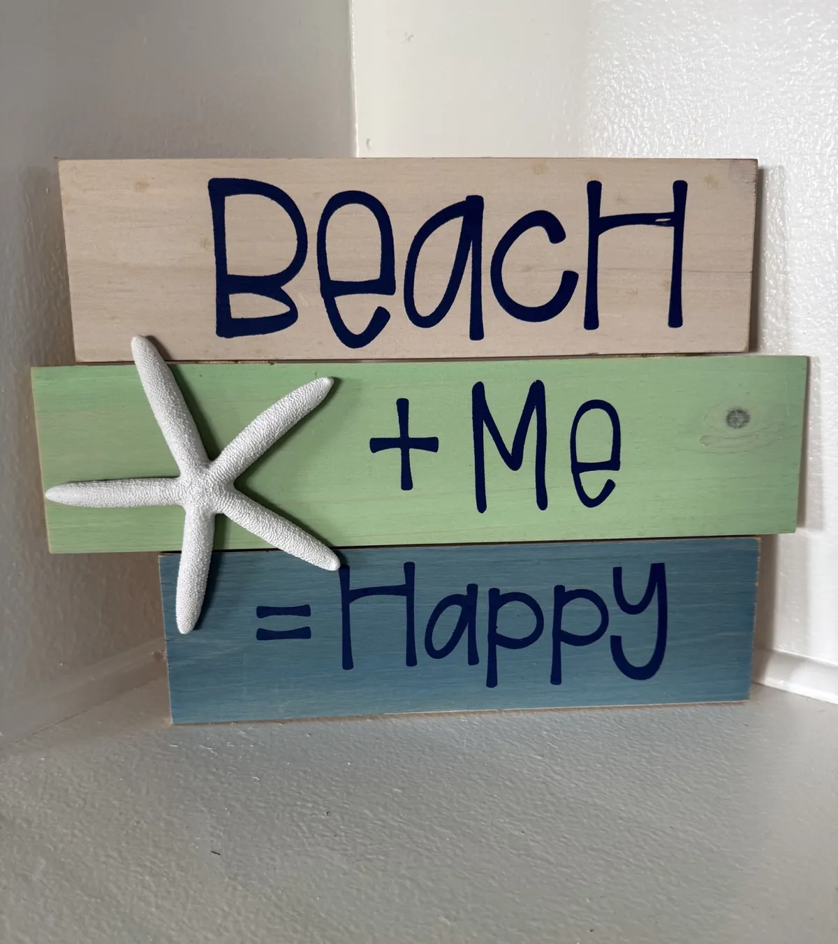 A close-up of a wooden beach-themed sign reading 'Beach + Me = Happy' with a white starfish accent, mounted on a light-colored wall. The décor is bright and uplifting, evoking a cheerful, coastal vibe that ties to the property's proximity to the beach and local attractions.