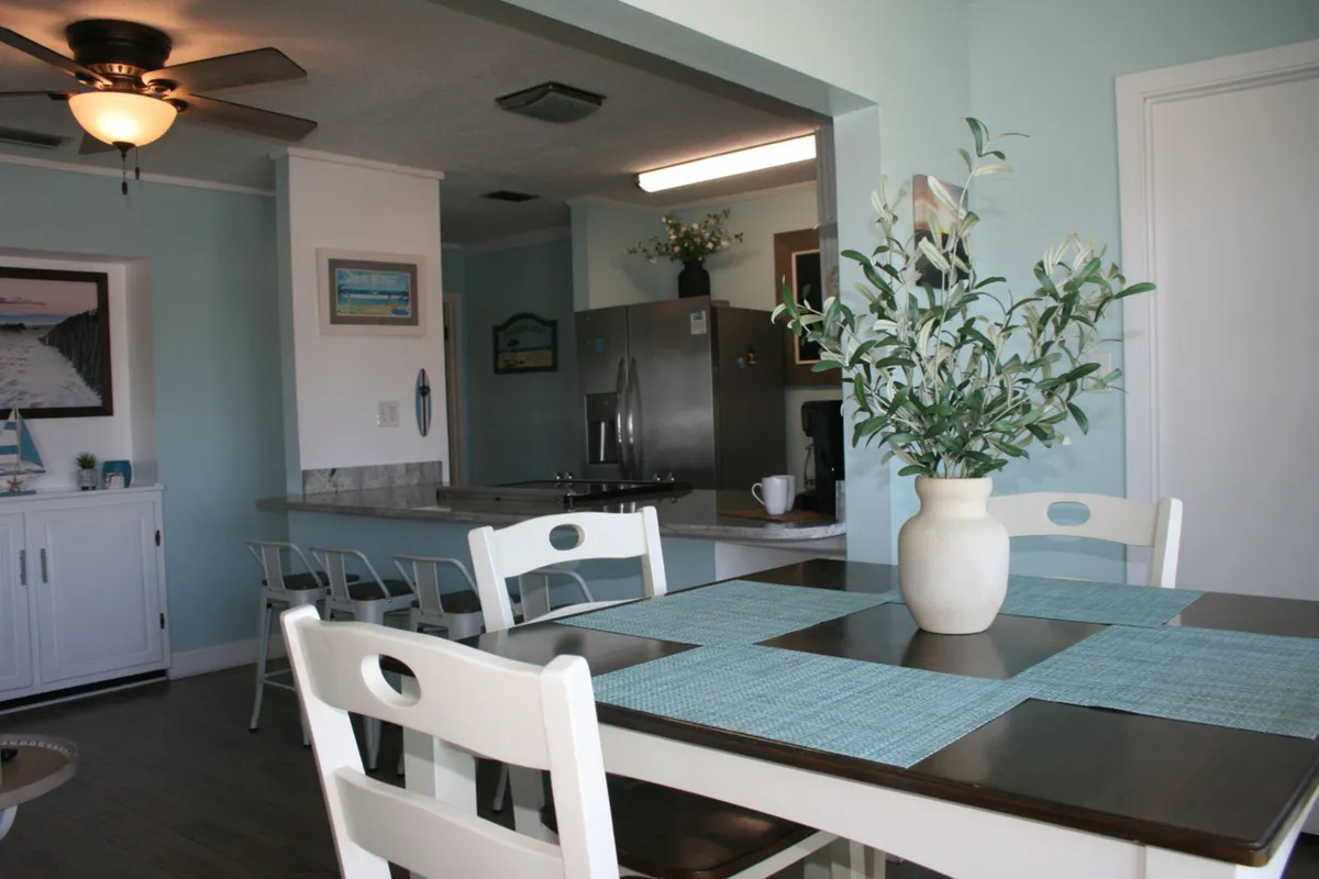 A dining area with a table set with blue placemats and a centerpiece vase of greenery sits adjacent to an open kitchen featuring stainless steel appliances, white cabinetry, and light blue walls. The room feels airy and calm, with a ceiling fan and natural light enhancing the space.