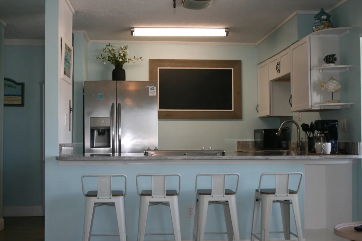 A bright, inviting kitchen area with a stainless steel refrigerator, a large blackboard with a wooden frame, and a kitchen counter lined with four white metal stools. The countertop holds small appliances, and the overall layout encourages group gatherings and casual meals.