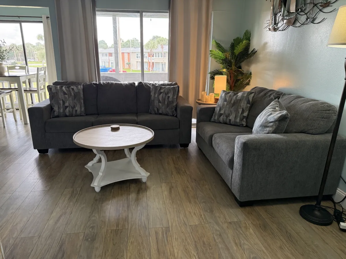 A bright, comfortable living room with two gray upholstered sofas adorned with patterned and solid throw pillows, a white round coffee table, and a large window letting in natural light and offering a glimpse of greenery and neighboring buildings. A tall potted plant and side table complete the inviting setting.