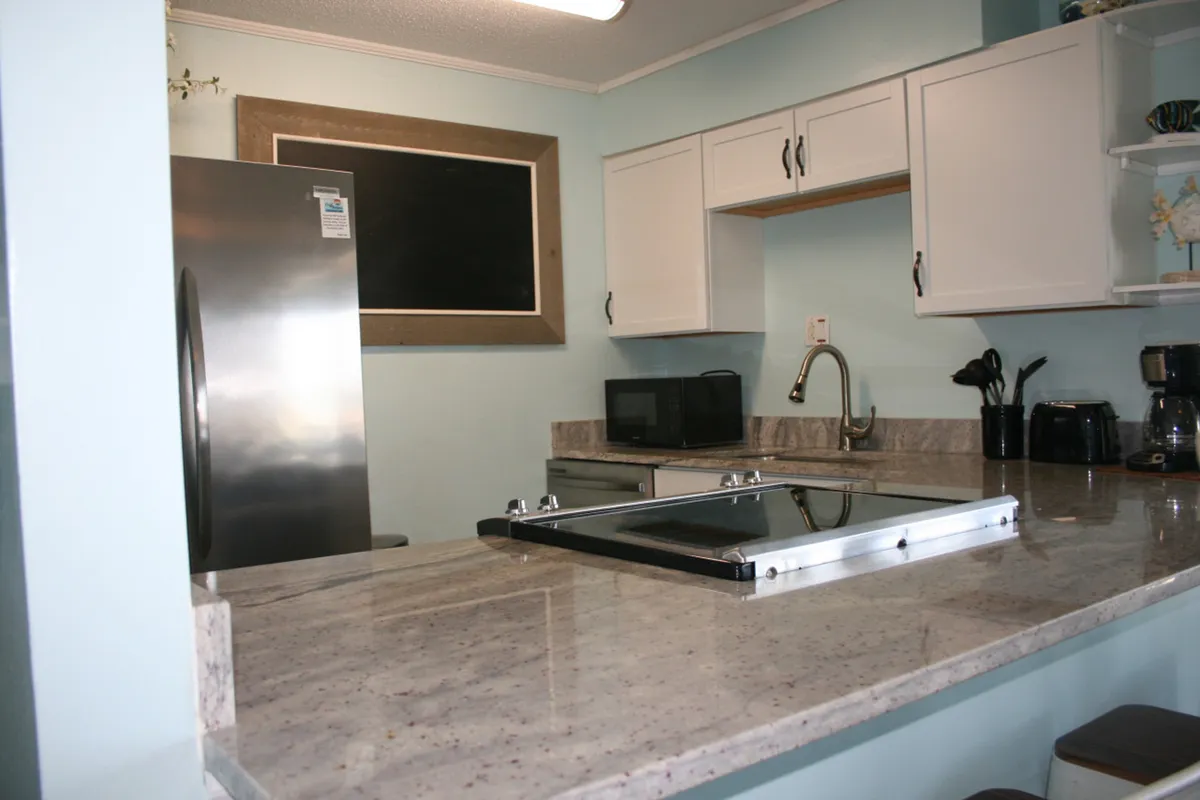 A modern kitchen featuring a large granite countertop with a built-in electric stove, stainless steel refrigerator, and white cabinetry with black handles. Black kitchen appliances, including a microwave, toaster, and coffee maker, are neatly arranged on the counters, creating a clean and practical cooking environment.