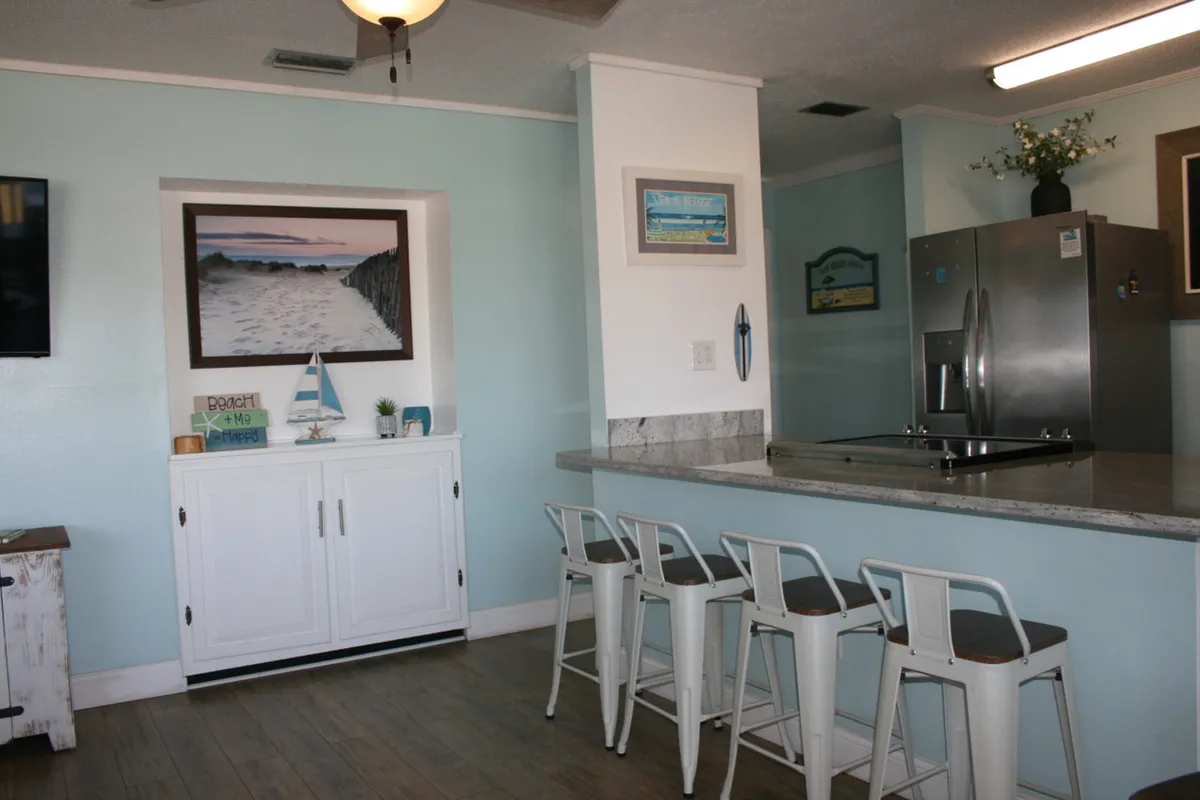 The kitchen island with four white bar stools is paired with a beach-themed cabinet and artwork, and a stainless steel refrigerator in the background. The setup encourages casual dining, conversation, and a sense of connection, all within a relaxed coastal-inspired interior.