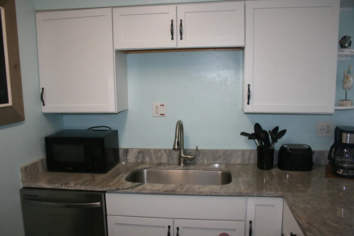 A close-up look at the kitchen sink area, featuring a stainless steel sink with a modern faucet, marble countertop, white cabinetry, and compact appliances like a microwave, toaster, and coffee maker. The space is clean and efficient, ideal for meal prep or quick cleanups.
