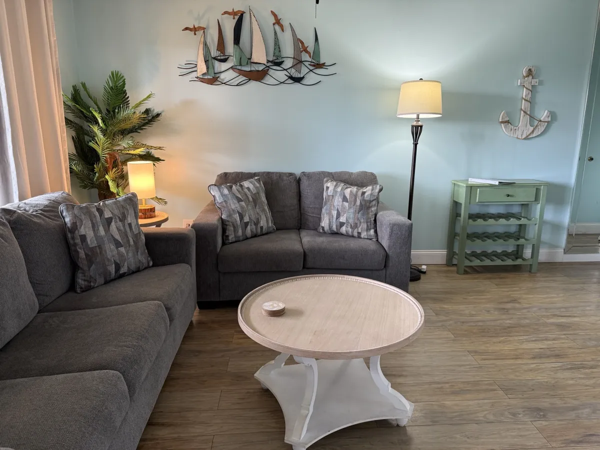 A spacious living area with a gray sectional couch adorned with patterned pillows, a round white coffee table, and nautical wall art featuring sailboats and birds. Warm lighting from a floor lamp adds to the inviting ambiance, suggesting a comfortable spot for relaxing after a day of activities.