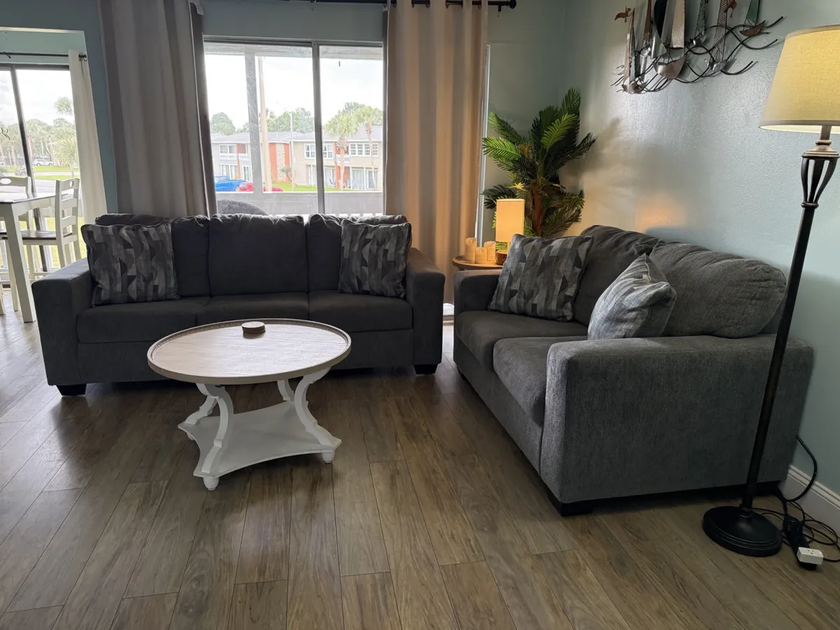 This image depicts a welcoming living area featuring two grey sofas arranged around a round white coffee table. A tall floor lamp, an indoor potted plant, and ample natural light contribute to a comfortable and inviting atmosphere ideal for relaxing or socializing.