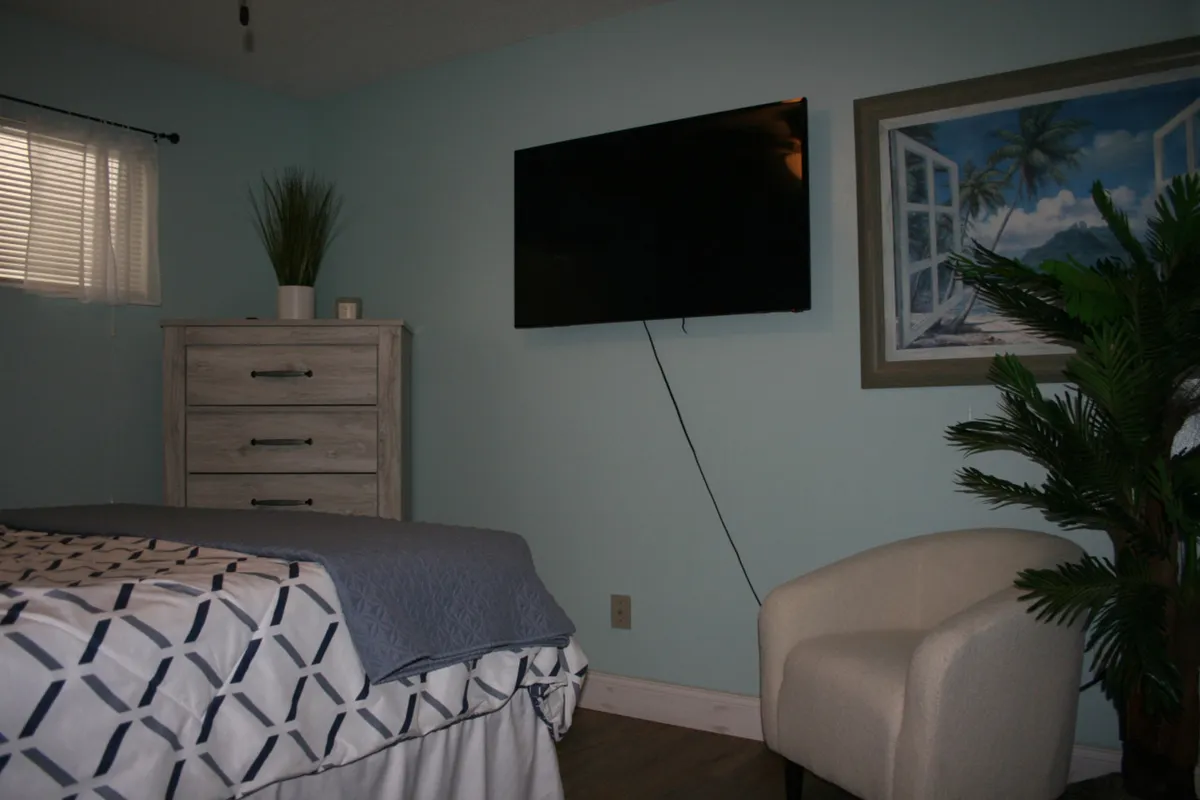 This bedroom scene features a patterned bedspread, wall-mounted flat screen TV, light wood dresser with a decorative plant, and framed tropical wall art. The lighting creates a cozy, restful mood ideal for unwinding.