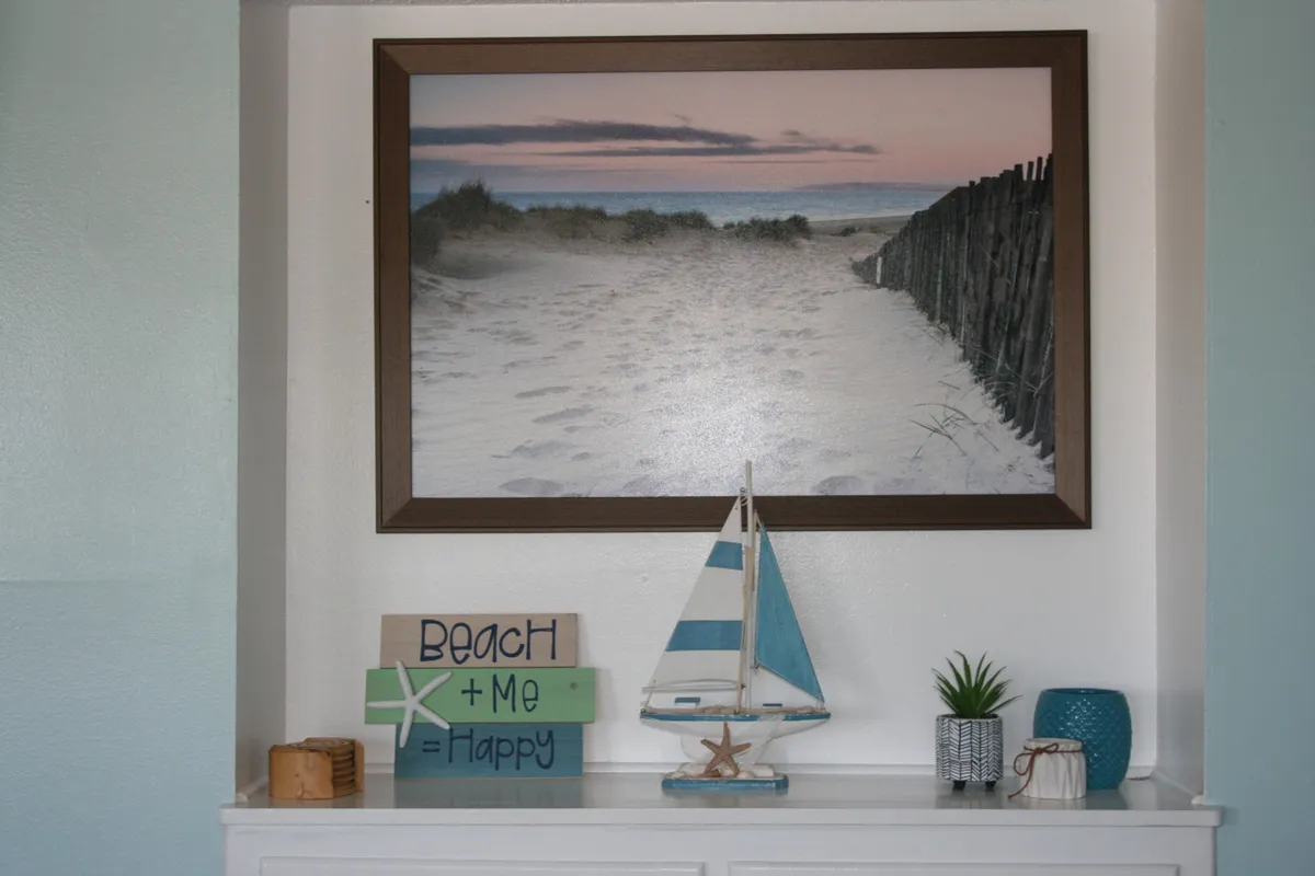 A cozy interior vignette featuring a framed beach photograph that depicts a sandy path to the ocean at either sunset or sunrise, a decorative wooden sign with the message 'Beach + Me = Happy', a small blue-and-white model sailboat, and small potted plants neatly arranged on a shelf. The decor is distinctly coastal, evoking a sense of being close to the beach and the outdoors.
