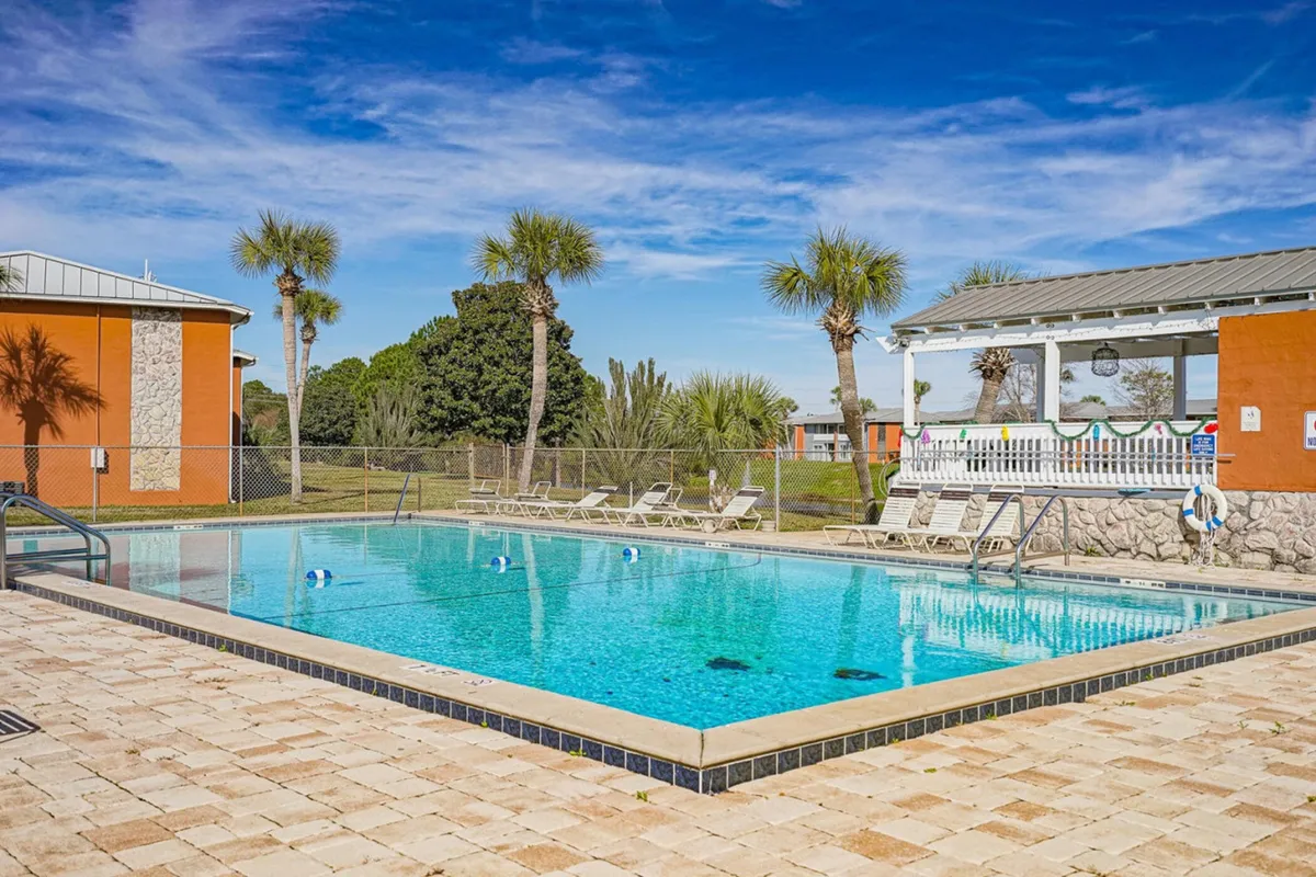 A vibrant scene featuring a sparkling community swimming pool, lounge chairs, and a covered seating area adorned with colorful decorations. Tall palm trees line the area, set beneath a bright blue sky, capturing the cheerful, leisurely atmosphere of the resort-style amenities. The space is designed for both swimming and casual outdoor socializing.