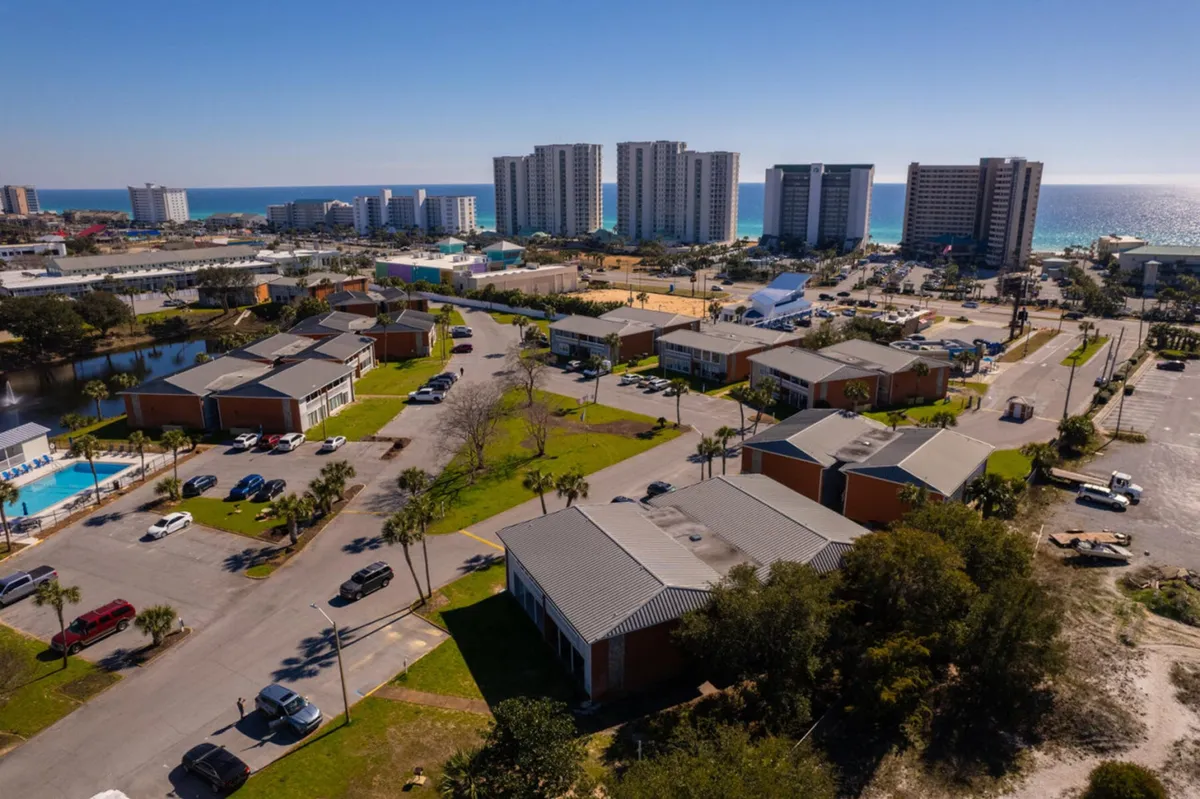 This exterior shot captures a cluster of low-rise residential buildings with visible parking, a bright blue swimming pool, and high-rise beachfront condos in the distance. The ocean horizon glimmers in the background, evoking a vibrant, coastal community environment. The image showcases both the pool amenities and the easy proximity to the beach and local attractions.