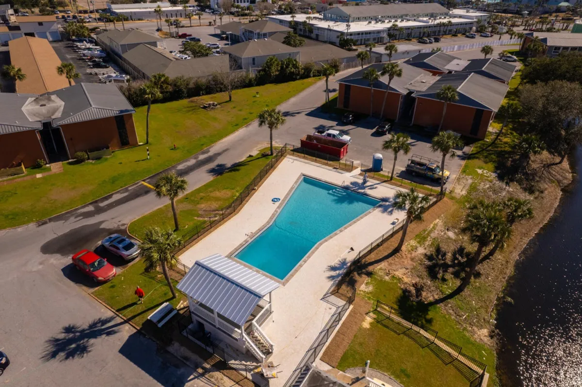 A rectangular outdoor swimming pool sits adjacent to a tranquil pond or lake with grassy gathering spaces and nearby residential buildings. Poolside shelter with seating offers a spot for picnics or family BBQs, while the calm water and greenery provide a peaceful, scenic backdrop.