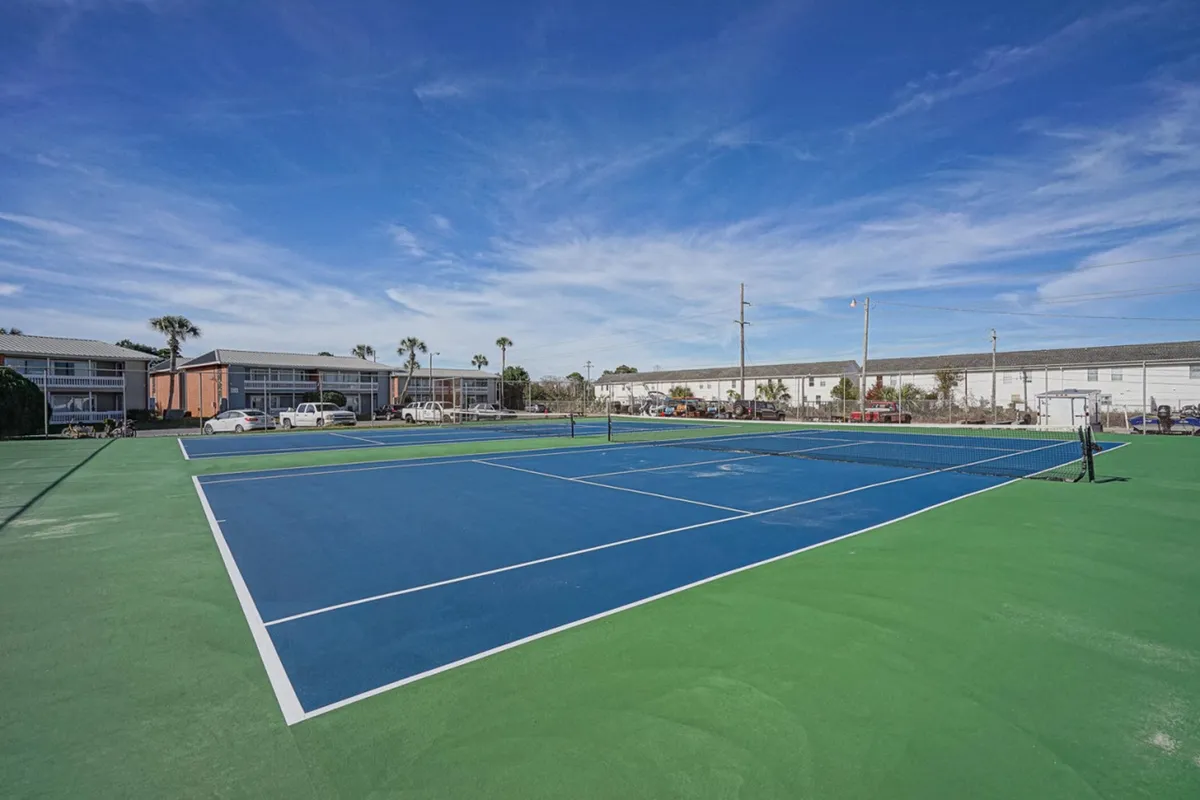 This image spotlights a well-maintained tennis court with a blue playing surface, surrounded by residential buildings and greenery. The clear blue sky and adjacent parking area emphasize the secure, active community atmosphere. The court is ready for play, inviting residents and guests to enjoy friendly matches or spectate.