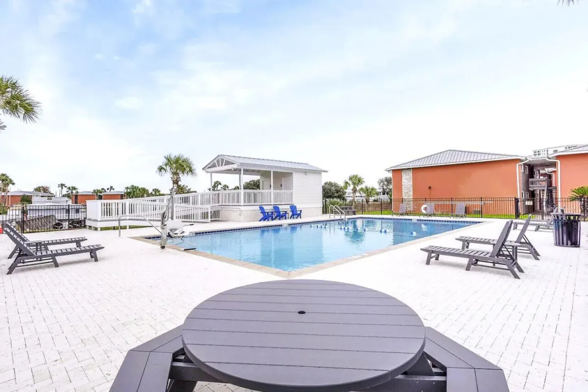 A bright, inviting swimming pool is the central focus, surrounded by poolside lounge chairs and a charming white gazebo. Vibrant orange buildings frame the backdrop, hinting at a lively community setting perfect for family gatherings and relaxation after a day at the water park.